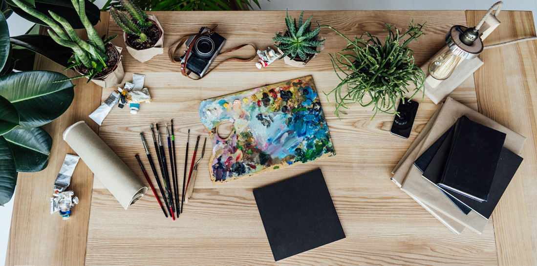 desk littered with creative objects: paintbrushes, plants, painting, paper